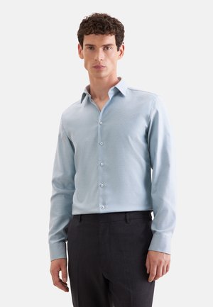 Mann mit lockigem Haar, der ein hellblaues langärmeliges Hemd und dunkle Hosen trägt, steht vor einem schlichten Hintergrund.