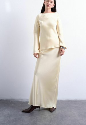 Femme portant un haut à manches longues en satin crème et une jupe longue assortie jusqu'au sol, associée à des bottes à talons pointues foncées et un bracelet argenté.