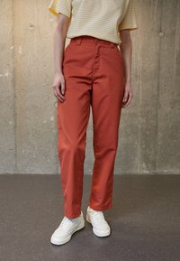 Roestige oranje rechte broek met een gladde textuur, voorzien van een sluiting met een knoop en ritssluiting aan de voorkant, gecombineerd met witte sneakers.