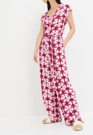 Femme portant une combinaison tie-dye rose et blanche à manches courtes avec une ceinture à la taille et des sandales blanches, debout devant un fond blanc uni.