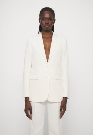 Donna con capelli intrecciati che indossa un blazer bianco sbottonato e pantaloni bianchi abbinati, in piedi contro uno sfondo chiaro e uniforme.