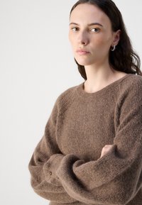Pulover texturat, de culoare maro, cu guler rotund și mâneci lungi, lejere, având detalii cu nasturi la manșete. Material moale și pufoasă.