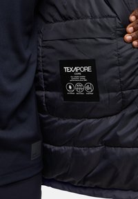 Giacca a vento nera con etichetta TEXAPORE, dotata di icone che indicano impermeabilità, traspirabilità e protezione dal vento. Texture morbida, trapuntata e design elegante.