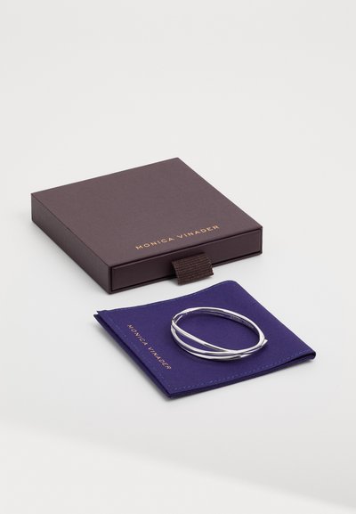 Bracelet en argent présenté sur un tissu bleu, à côté d'une boîte violet foncé avec du texte doré et une fermeture à ruban marron.