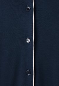 Námořnický modrý cardigan vyrobený z texturované látky, který má šest kontrastních knoflíků a bílou lemovku podél předního otvoru.