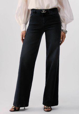 Donna che indossa jeans neri a vita alta a gamba larga, camicetta bianca con maniche a sbuffo, tacchi neri open toe con dettaglio a catena dorata.