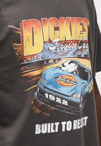 Γκρι βαμβακερό μπλουζάκι με πολύχρωμο γραφικό ενός αγωνιστικού αυτοκινήτου, το λογότυπο "Dickies" και το κείμενο "BUILT TO RESIST" με έντονα λευκά γράμματα.
