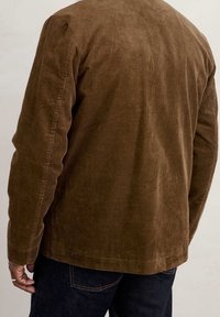 Brązowa kurtka w korduroy z prążkowanym wzorem, z długimi rękawami i prostym krojem, noszona na ciemnych dżinsach, o prostym designie.