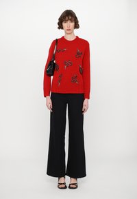 Red knitted sweater with snake embellishments, paired with black wide-leg pants and black sandals. Model holds a small black handbag.