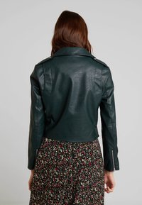 Grønne faux læder motorcykeljakke med et cropped design, bred krave og lynlåse ved manchetterne, parret med en blomstret kjole.