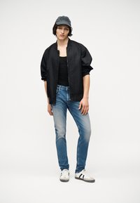 Νέος άνθρωπος φορώντας σκούρο μπουφάν τύπου bomber, μαύρο αμάνικο μπλουζάκι, μπλε τζιν, γκρι καπέλο και λευκά αθλητικά παπούτσια, στέκεται μπροστά σε λευκό απλό φόντο.