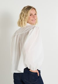 Blusa blanca de algodón con mangas abullonadas y detalle fruncido en la parte posterior, combinada con jeans de mezclilla oscuros. Textura suave y corte relajado.