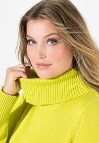 Maglione lavorato a maglia di un vivace verde lime con un colletto alto a costine. In primo piano si vede la texture dettagliata e la vestibilità, con le mani che afferrano parzialmente il colletto.