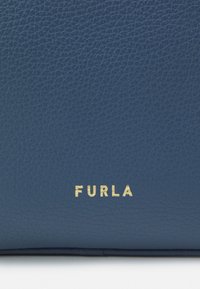 Marinblå läderhandväska med strukturerad yta. Har en guldprickad logotyp "FURLA" längst ner som anger varumärkesidentitet.