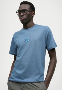 T-shirt de algodão azul claro com mangas curtas, apresentando um logo da Rip Curl em tons de azul escuro e verde no centro. Design de ajuste descontraído.