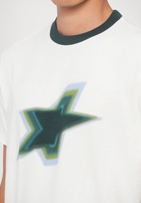 Biała bawełniana koszulka z grafiką rozmytej gwiazdy w odcieniach zieleni i niebieskiego, z ciemnozielonym prążkowanym kołnierzykiem i gładką teksturą.