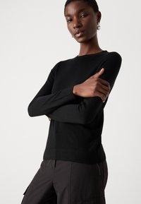 Μαύρο πλεκτό πουλόβερ με μακριά μανίκια, στρογγυλή λαιμόκοψη, λεία υφή, εφαρμοστό σχέδιο, φορεμένο με σκούρες cargo παντελόνες.