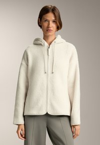 Giacca di pile color crema con cappuccio, zip frontale e cordini. Presenta una superficie testurizzata e una vestibilità rilassata per il massimo comfort.