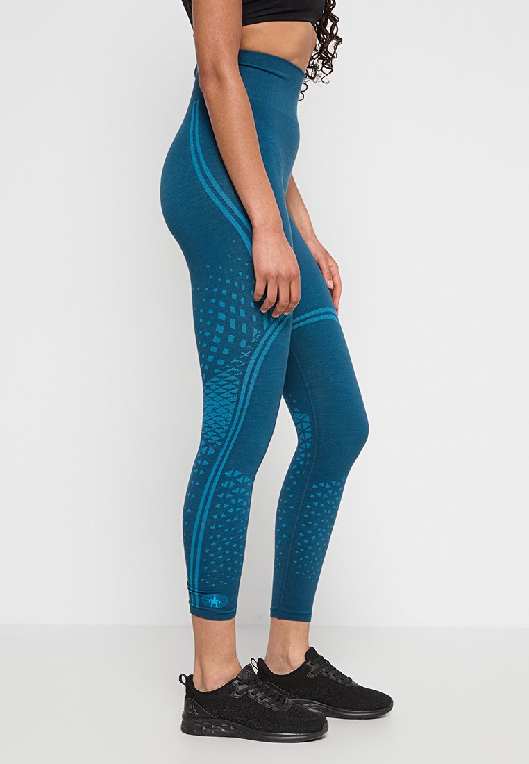 Kobieta ubrana w tealowe legginsy z wysokim stanem w wzory oraz czarne buty sportowe, stojąca bokiem na jednolitym, jasnym tle.