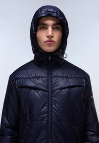 Giacca a vento blu navy con colletto alto e cappuccio regolabile, dotata di chiusura a zip, tasche laterali e una patch rotonda con logo sul braccio.