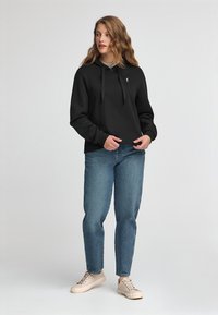 Felpa nera con cappuccio e lacci, logo sul davanti, abbinata a jeans blu e sneakers beige. Tessuto liscio con una vestibilità comoda.