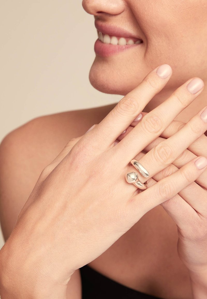 Deux anneaux en argent sur des doigts, l'un avec un design lisse et arrondi et l'autre avec une forme bulbeuse, sur un fond neutre.