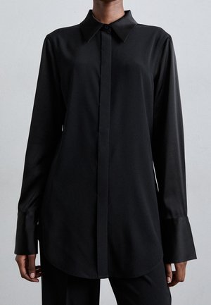 Person trägt ein langärmliges, schwarzes Hemd mit Knöpfen, spitzem Kragen und breiten Manschetten vor einem einfarbigen Hintergrund.