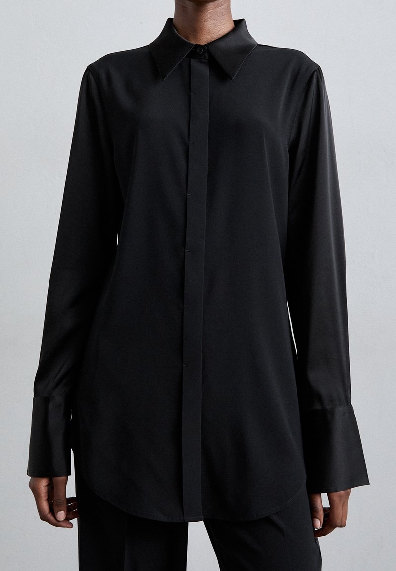 Osoba ubrana w czarną koszulę z długim rękawem, zapinaną na guziki, z ostrym kołnierzykiem i szerokimi mankietami na jednolitym tle.