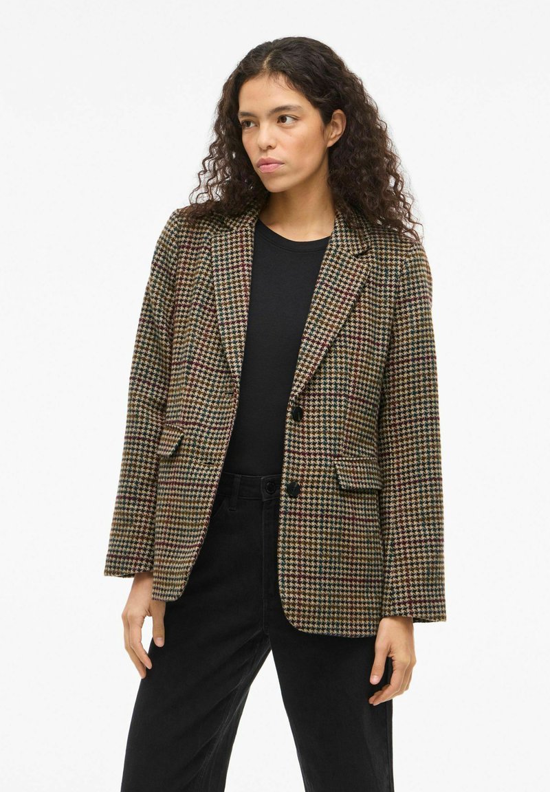 Houndstooth-patterned blazer in brown, green, and red with a tailored fit, notched lapels, and two front pockets, worn over a black top.