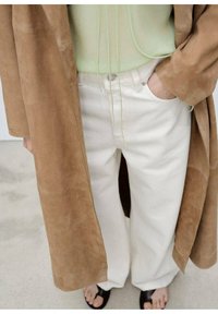 Hnědý semišový kabát přes světle zelený top, doplněný bílými kalhotami s širokými nohavicemi. Černé sandály dotvářejí outfit. Hladká textura a čisté linie.
