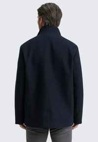 Palton din lână de culoare bleumarin, cu guler înalt, umeri structurați și croială dreaptă. Prezintă o cusătură minimă și o textură netedă.