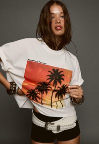 White t-shirt with a colorful sunset graphic featuring palm trees, paired with black shorts and a studded white belt.
