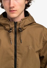 Chaqueta con capucha marrón que presenta un cierre con cremallera, cordones ajustables, forro negro y un logotipo bordado en el pecho.
