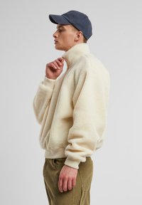 Kremowy polarowy sweter z wysokim kołnierzem, prążkowanymi mankietami i dołem, z zamkiem błyskawicznym. Połączony z oliwkowymi spodniami sztruksowymi i granatową czapką.