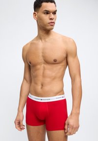 Oberkörperfreier Mann trägt rote Tommy Hilfiger Boxershorts mit weißem Bund, steht vor einem schlichten hellen Hintergrund und schaut zur Seite.