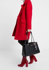 Sac à main noir avec poignées en chaîne, texture brillante et pendentifs décoratifs. Manteau rouge avec une finition texturée et des manches longues. Bottes à talons hauts rouges.