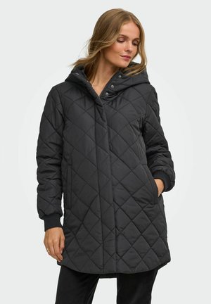Žena nosi čierny prešívaný kabát s kapucňou, prednými vreckami, dlhými rukávmi a zapínaním na zips a patentné gombíky, pozerá sa nadol do strany.