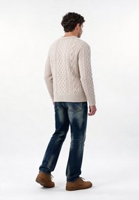 Homme portant un pull en maille torsadée beige, un jean bleu délavé et des chaussures marron à lacets, debout de dos sur un fond blanc.