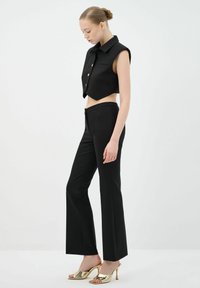 Black cropped vest with a collar and gold buttons paired with high-waisted black trousers, featuring a sleek fit and flared legs. Gold heeled sandals.
