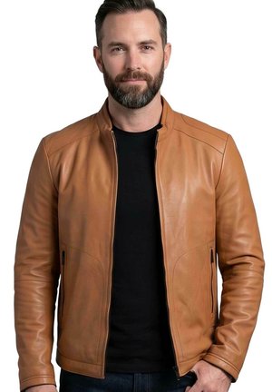 Man with short dark hair and beard wearing a tan leather jacket zipped open over a black shirt, standing against a white background.