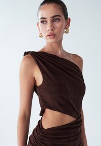 Vestito marrone monospalla con tessuto drappeggiato, spacco laterale e dettagli arricciati. Il materiale testurizzato aumenta l'interesse visivo. Orecchini dorati completano il look.