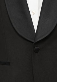 Gros plan sur une veste de smoking noire avec revers en satin, bouton unique recouvert de tissu, associée à une chemise blanche et un bouton blanc.