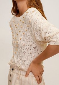 Sweter dziergany w kolorze off-white z teksturowanym wzorem, z wypukłymi koralikami wzdłuż dekoltu i górnej części klatki piersiowej, oraz luźnymi rękawami.