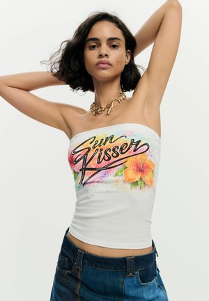 Jeune femme aux cheveux courts et foncés portant un haut blanc sans bretelles avec un texte coloré « Sun Kisser », un imprimé floral, un collier doré et un jean bleu.