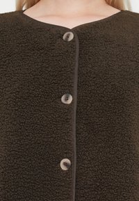 Brown fleece garment with a textured surface, rounded neckline, and button closure featuring a cream-colored button at the center.