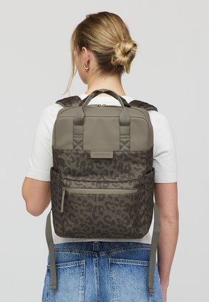 Frau mit blondem Haarknoten, die ein weißes Hemd und blaue Jeans trägt, trägt einen grauen Rucksack mit Leopardenmuster und Frontreißverschlusstasche.