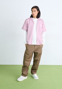 Camicia rosa a maniche corte con bottoni sopra una maglietta bianca a maniche lunghe, abbinata a pantaloni larghi marroni e sneaker bianche su un pavimento verde.