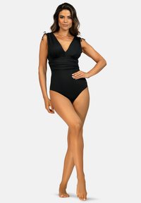 Kobieta stoi boso, jedna ręka na biodrze, ubrana w czarny jednoczęściowy kostium kąpielowy bez rękawów z głębokim dekoltem w kształcie litery V i marszczonym pasem.