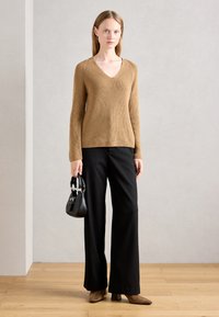 Pull en maille côtelée marron avec décolleté en V, associé à un pantalon noir à jambes larges. Le mannequin tient un petit sac à main noir et porte des bottes à talons marron.