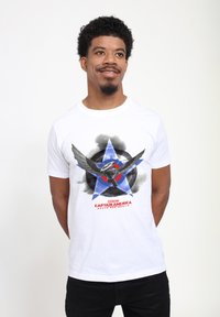 Camiseta de algodón blanca con un gráfico de un personaje en vuelo, contra un fondo de estrellas azules y nubes negras, con letras rojas.